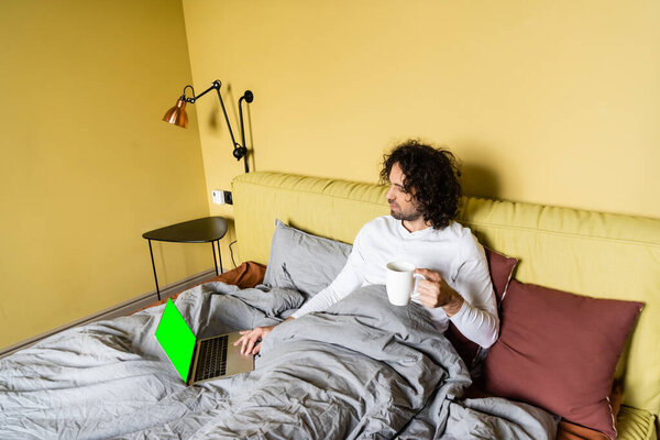 high angle view of young freelancer using laptop with green screen while holding cup of coffee in bed