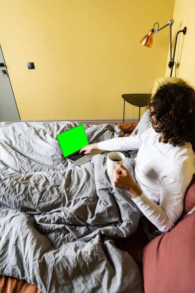 high angle view of curly freelancer using laptop with green screen while holding cup of coffee in bed