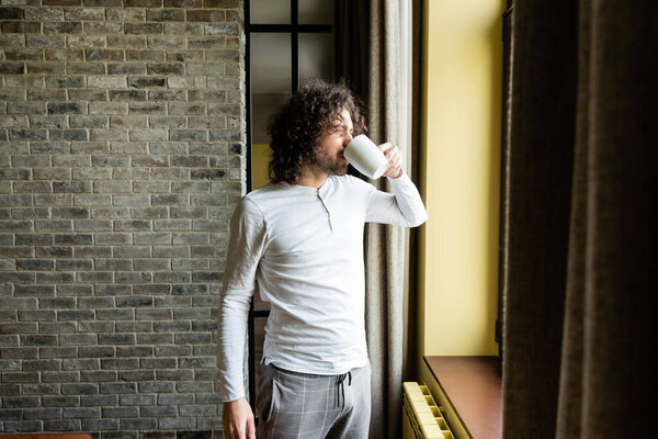 Красивый мужчина в пижаме пьет кофе, глядя в окно утром
