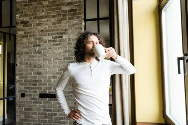 handsome man in pajamas drinking coffee while standing with hand on hip and looking at camera