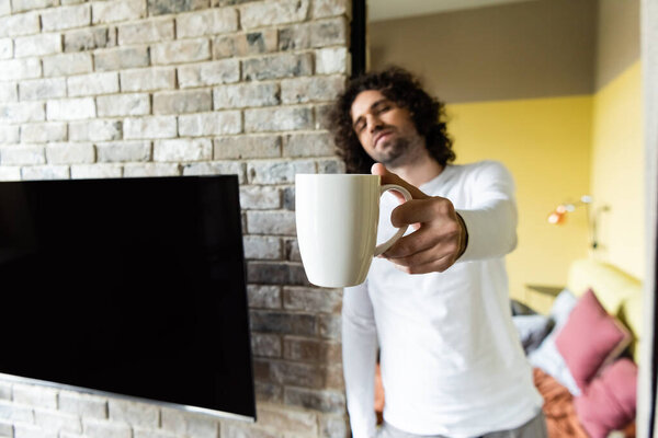 selective focus of handsome man holding coffee cup in outstretched hand near blank lcd screen hanging on brick wall