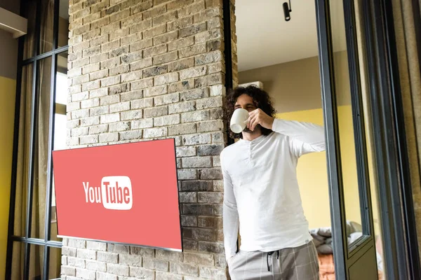 stock image KYIV, UKRAINE - APRIL 25, 2020: young man in pajamas drinking coffee while standing near lcd screen with Youtube website