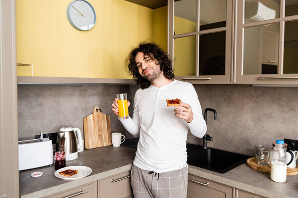 happy man in pajamas holding orange juice and toast with jam while smiling at camera