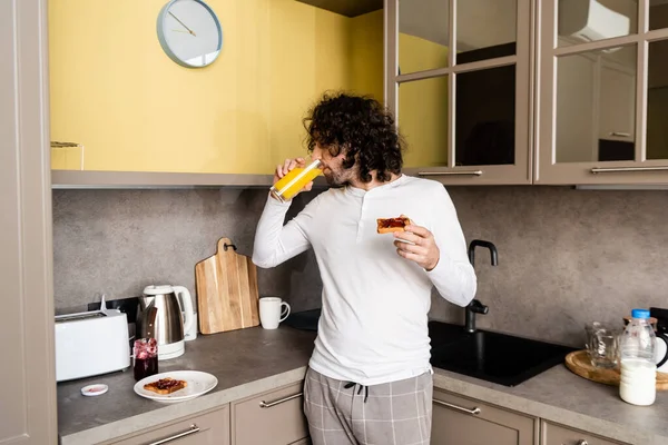 young man in pajamas drinking orange juice and holding toast with jam in kitchen