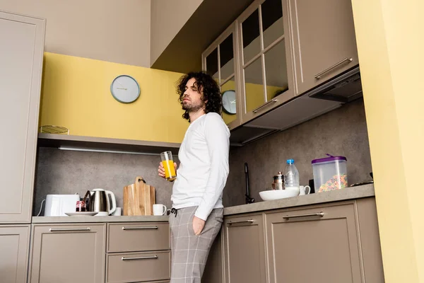 thoughtful man in pajamas standing with hand in pocket while holding orange juice in kitchen