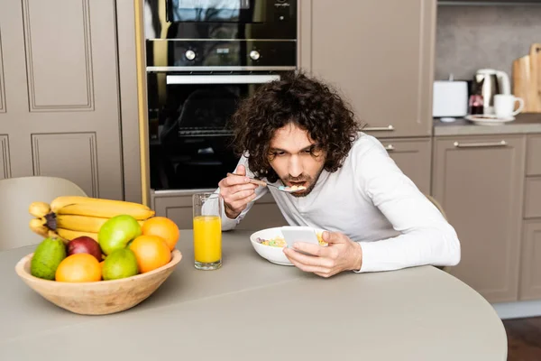 Concentrated Curly Man Chatting Smartphone Breakfast Kitchen Bowl Fruits — Stock Photo, Image