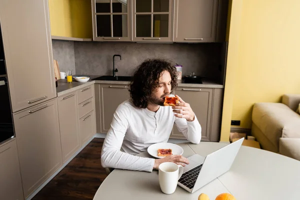 Handsome Freelancer Eating Toast While Using Laptop Coffee Cup Kitchen — Stock Photo, Image