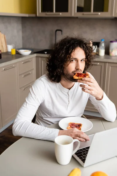 Guapo Freelancer Comer Tostadas Mirando Cámara Cerca Portátil Taza Café — Foto de Stock