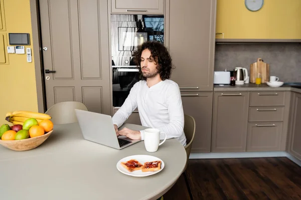 back view of freelancer using laptop with web shopping website on screen near coffee cup, toasts and fresh fruits