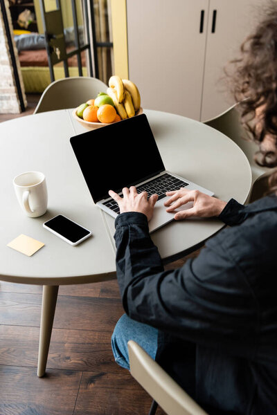 back view of freelancer using laptop with blank screen near smartphone, coffee cup, sticky notes and fruits