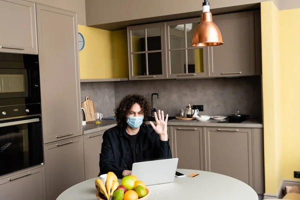 戴着医疗面罩的男人 在智能手机旁边的笔记本电脑上有视频通话 在厨房的桌上有信用卡 — 图库照片