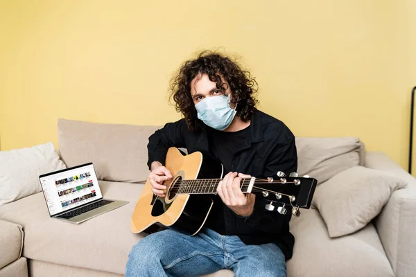KYIV, UKRAINE - 25 Nisan 2020: Medikal maskeli adam dizüstü bilgisayarın yanında akustik gitar çalıyor 