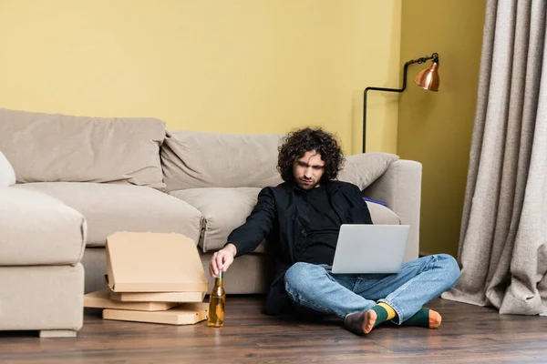 Freelancer holding bottle of beer near laptop and pizza boxes on floor in living room