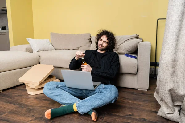 Freelancer opening bottle of beer near laptop and pizza boxes on floor