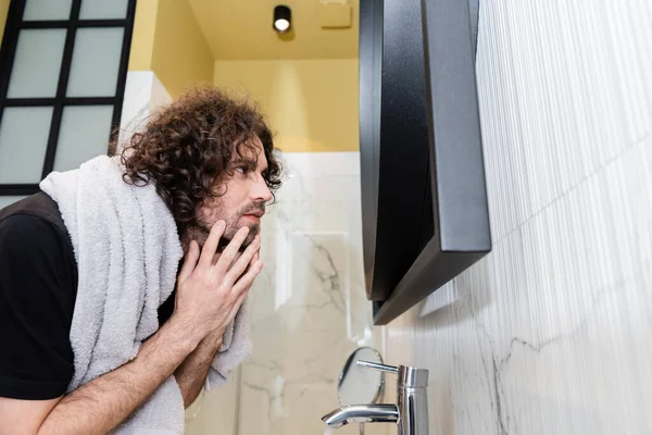 Side view of handsome curly man touching face while looking at mirror in bathroom
