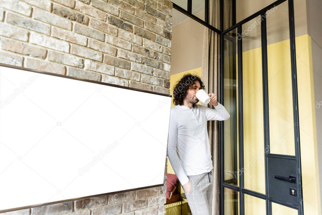 young man in pajamas drinking coffee while standing near white lcd screen hanging on brick wall