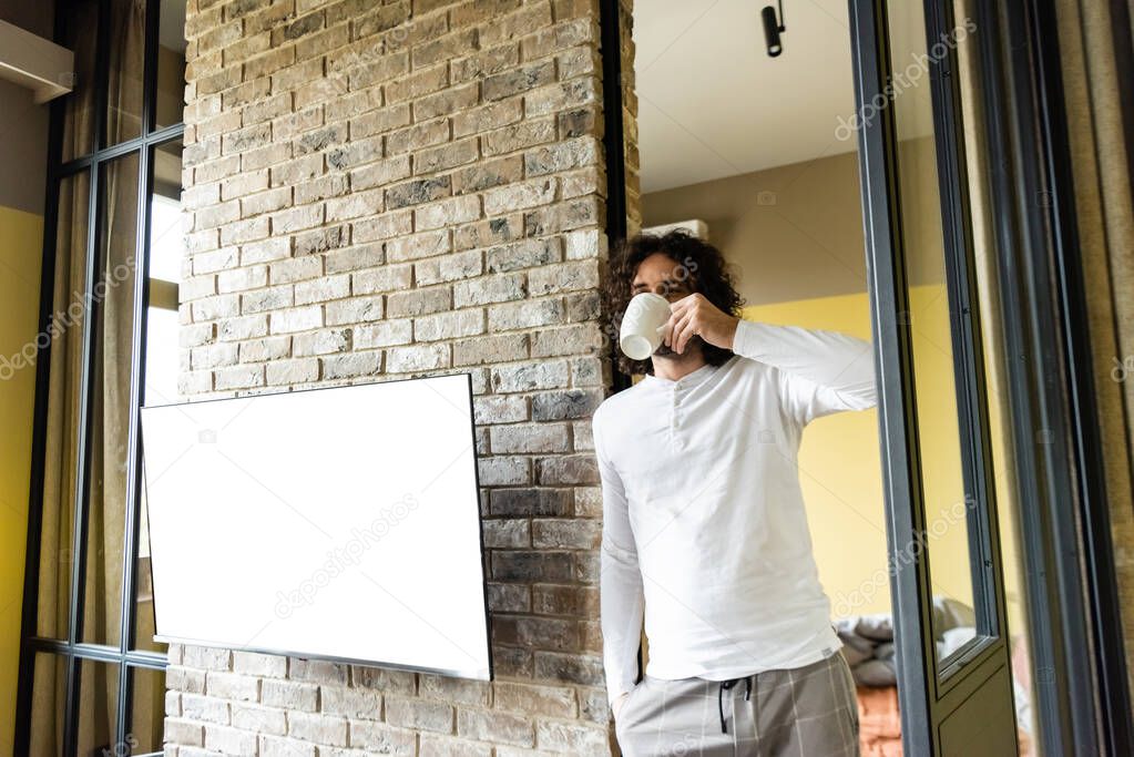 young man in pajamas drinking coffee while standing near white lcd screen hanging on brick wall
