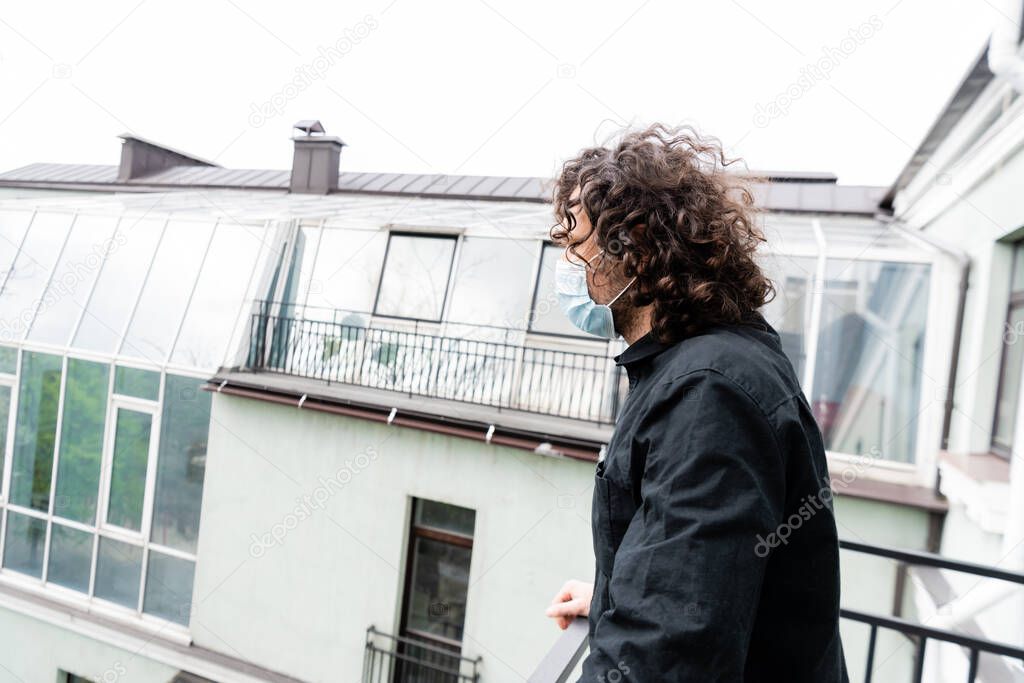 Side view of curly man in medical mask standing on balcony at home 