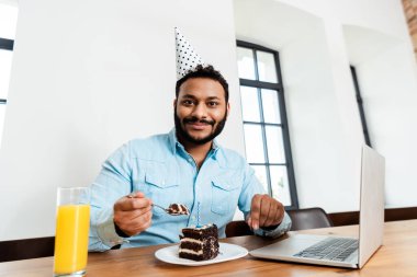 Mutlu Afrikalı Amerikalı serbest parti şapkası takmış, elinde lezzetli doğum günü pastası ve dizüstü bilgisayarının yanında kaşık tutuyor. 