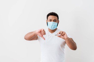 Tıbbi maskeli hasta Afro-Amerikan adam dijital termometre tutuyor ve beyaz duvarın yanında baş parmağını gösteriyor. 