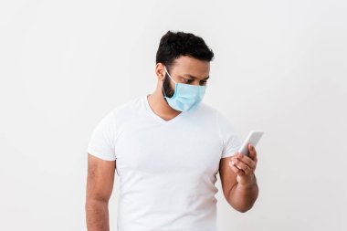 Beyaz duvarın yanında akıllı telefon kullanan tıbbi maskeli Afro-Amerikan bir adam. 