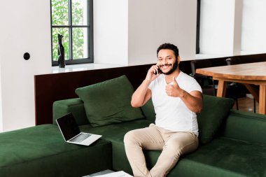 Sakallı Afrikalı Amerikalı adam akıllı telefondan konuşuyor ve dizüstü bilgisayarın yanında boş ekranla baş parmağını gösteriyor.