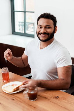 Kavanozun yanında tatlı reçelli kaşık ve tabakta kızarmış ekmek tutan mutlu Afrikalı Amerikalı adam.