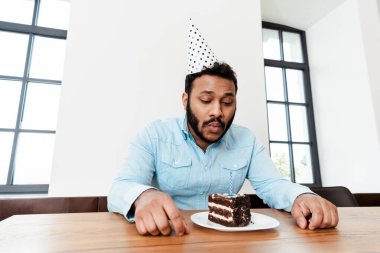 Parti şapkalı Afro-Amerikan adam doğum gününü yalnız kutluyor ve doğum günü pastasında mum üflüyor.