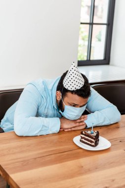 Parti şapkalı ve tıbbi maskeli Afro-Amerikan adam doğum gününü yalnız kutluyor ve doğum günü pastasına bakıyor.