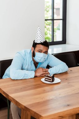 Parti şapkası ve sağlık maskesi takmış Afro-Amerikan bir adam doğum gününü lezzetli doğum günü pastasının yanında tek başına kutluyor.