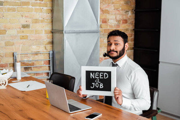 bearded african american man holding chalkboard with need a job lettering near gadgets 