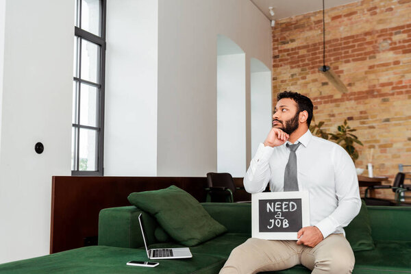 upset african american man holding chalkboard with need a job lettering near gadgets on sofa