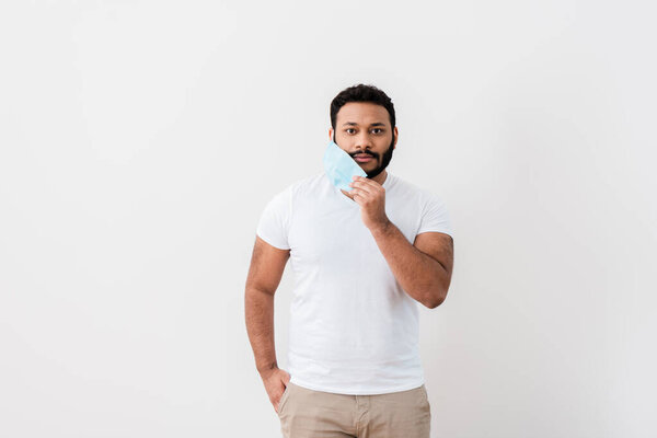 бородатый африканский американец трогает синюю медицинскую маску и стоит с рукой в кармане возле белой стены
 