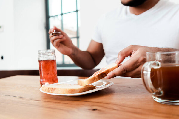 cropped view of african american man holding spoon near jar with sweet jam and toast bread on plate