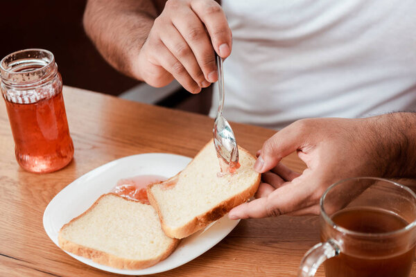 cropped view of african american man holding spoon with sweet jam near toast bread on plate