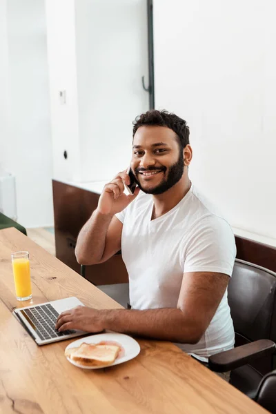 快乐的非洲裔美国自由职业者 一边在笔记本电脑旁一边用智能手机聊天 一边在桌上吃早餐 — 图库照片