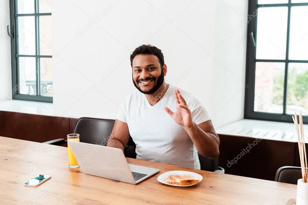 happy african american man waving hand while having video chat near tasty breakfast on table