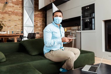Tıbbi maskeli ve parti şapkalı Afro-Amerikan adam oturma odasındaki laptopun yanında lezzetli bir doğum günü pastası tutuyor.