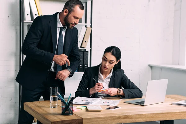 Hombre de negocios apuntando a la guardia cerca de la mujer de negocios concentrada en el lugar de trabajo con computadora portátil en la oficina - foto de stock