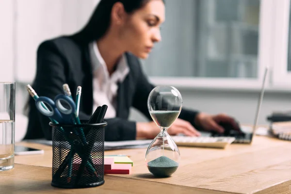 Selective focus of hourglass and businesswoman in suit working on laptop at workplace in office — Stock Photo