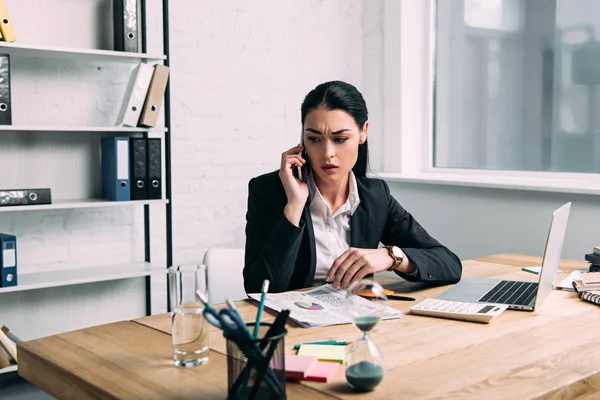 Femme d'affaires en costume parler sur smartphone sur le lieu de travail avec calculatrice et papiers au bureau — Photo de stock