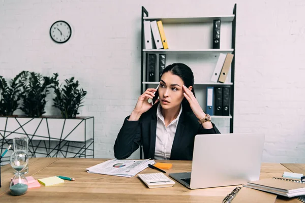 Mujer de negocios en traje hablando en smartphone en el lugar de trabajo con calculadora y papeles en la oficina - foto de stock