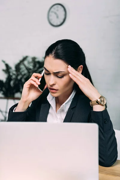 Stressed businesswoman in suit talking on smartphone at workplace with laptop in office — Stock Photo