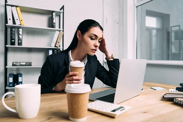 Tired businesswoman in suit with coffee to go working on laptop at workplace in office — Stock Photo