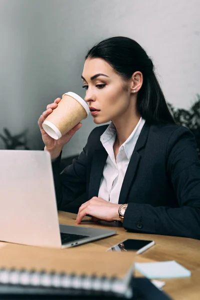 Side view of businesswoman in suit with disposable cup of coffee at workplace with laptop in office — Stock Photo