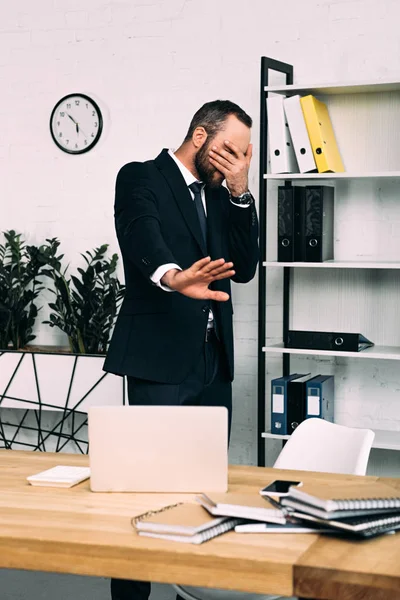 Vue obscurcie de l'homme d'affaires fatigué couvrant le visage avec la main sur le lieu de travail en costume au bureau — Photo de stock