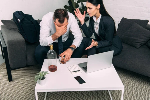 Vista parcial de los colegas de negocios estresados en la mesa de café con computadora portátil y alcohol en la oficina - foto de stock