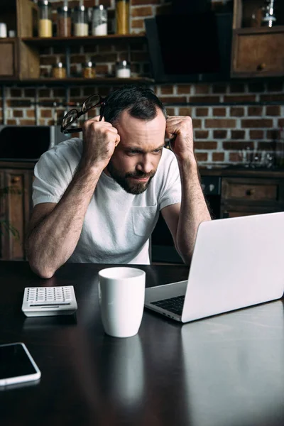 Retrato del freelancer sobrecargado trabajando a distancia en la mesa con el ordenador portátil en la cocina en casa - foto de stock