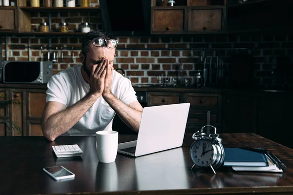 Freelancer sobrecargado trabajando a distancia en la mesa con el ordenador portátil en la cocina en casa - foto de stock