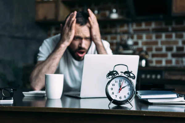 Enfoque selectivo de freelancer estresado a distancia trabajando en la mesa con el ordenador portátil y el reloj en la cocina en casa - foto de stock